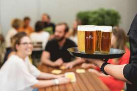 Bei den Ottakringer Braukultur-Wochen freut sich eine Gruppe am Tisch auf die Ottakringer Biere, die im Vordergrund auf einem Tablett herangetragen werden