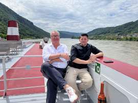 Tim Mlzer mit DDSG-Blue-Danube-Geschftsfhrer Wolfgang Fischer sitzen am Oberdeck eines Schiffes, im Hintergrund die Wachau