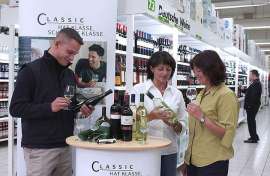 Kunden im LEH verksoten Wein an einem Stand vor vollen Regalen mit Deutschem Wein