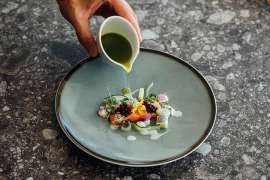 Gourmetkche im Falkensteiner Balance Hotel Stegersbach: wunderschn angrichtetes Lachfilet mit Blten und Beeren aus dem Permagarten, ein grnes Kruterl wird angegossen