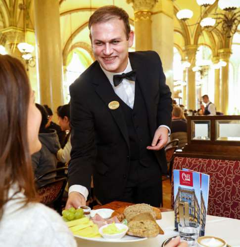 Ein lächelnder Ober serviert einer Dame Frühstück im Café Central