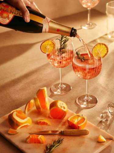 zwei hübsch dekorierte Gläser in die aus der Flasche Chandon Garden Spritz eingeschenkt wird, davor ein Schneidbrett mit Messer und Orangenscheiben