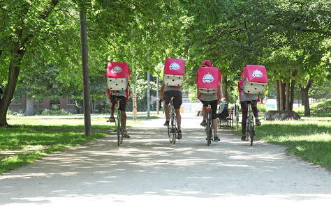 4 FAhrradboten mit rosa Foodora-Rucksäcken fahren in einem Park in Blickrichtung davon