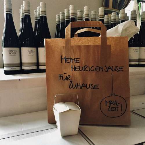 Heurigen to Go - vor einigen Weissweinflaschen steht die Heurigenjause liebevoll in ein Papiersackerl verpackt mit der Aufschrift 'Meine Heurigenjause - für Zuhause Mahlzeit!'