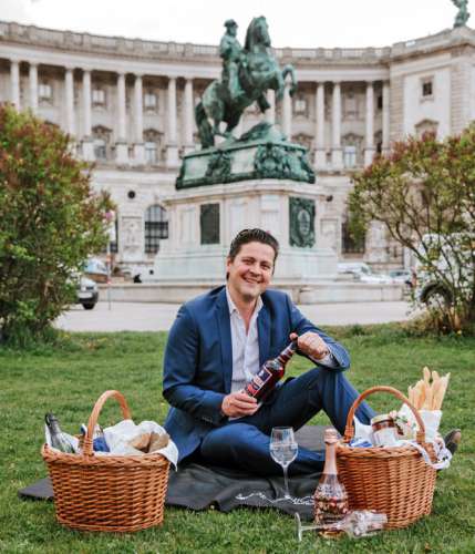 Paul Rittenauer auf einer Decke in der Wiese vor dem Reiterdenkmal am Heldenplatz, vor ihm 2 Picknickkörbe, in der Hand eine Flasche Spritz Select